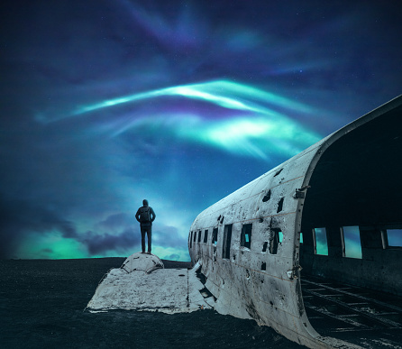 Man standing on a wing of a plane wreck on the black beach at Solheimasandur in the South coast of Iceland under the northern lights.