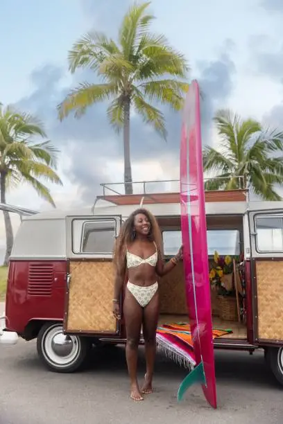 A tropical shot of a black woman with a surfboard next to a Volkswagen van on the beach in Hawaii