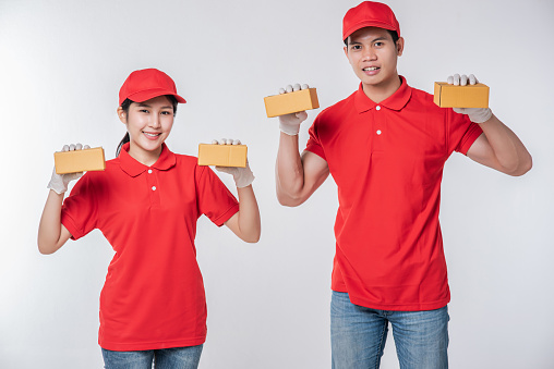 Image of a happy young delivery man in red cap blank t-shirt uniform standing with empty brown cardboard box isolated on light gray background studio