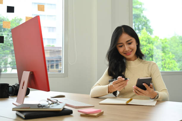 istock Charming young female freelancer sitting front of computer pc and using mobile phone at home office. 1437192269