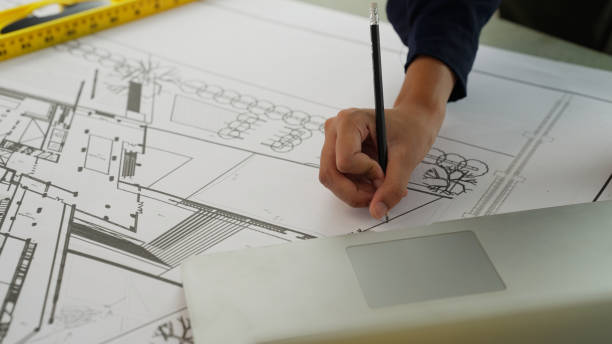 Hardworking female architect with yellow helmet drawing on blueprint of architectural project plan on building site using computer laptop working on high building, Construction work concept
