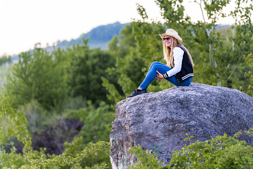A shallow focus shot of a European female with cowboy hat sitting on a rock in nature