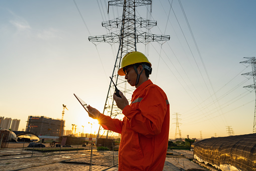 Power engineer using tablet to record checking power transmission line at sunset