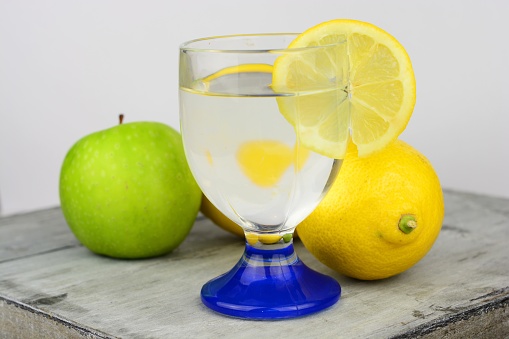 A closeup shot of a glass of lemon juice with a sliced lemon next to whole lemons and an apple