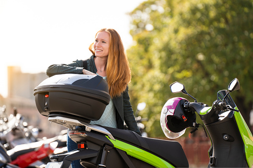 Portrait of a red-haired woman on her electric motorcycle in a parking lot talking on smartphone