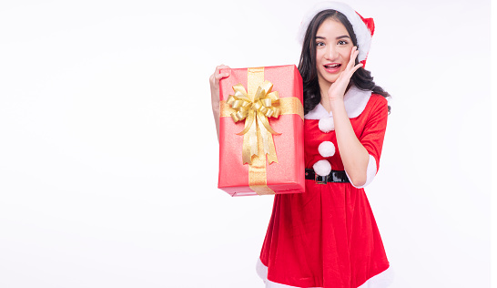 Excited asian woman wear SantaClaus dress holding red gift box standing on white background. Beautiful Santa Claus lady with hat holding hand beside mouth surprise gift box over isolated background.