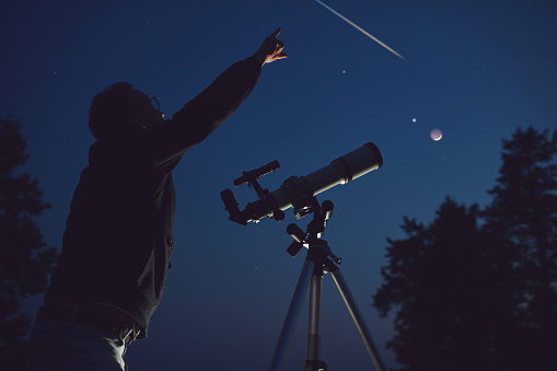 Silhouette of a man, telescope, stars, planets and shooting star under the night sky.
