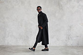 Fashionable man wearing black coat, turtleneck, pants, loafers shoes, glasses in studio. Fashion african american male model in stylish clothes, total black