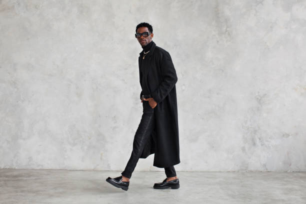 Fashionable man wearing black coat, turtleneck, pants, loafers shoes, glasses in studio. Fashion african american male model in stylish clothes, total black stock photo