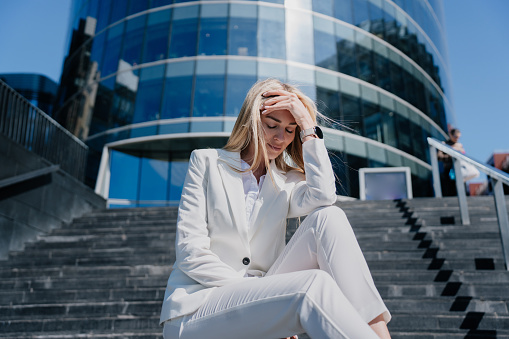 Young blonde Italian woman in white suit sit-in on chairs in despair against huge business center, employee loosed her job, troubles. Upset, disappointed woman eyes closed touching head, frustrated.