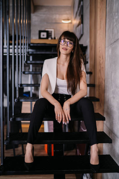 istock Young Italian brunette with loose hair dressed elegantly sitting on the steps in office, needs a brake. Vertical shot of beautiful caucasian girl in glasses exhausted after work. Lawyer at office 1437225464