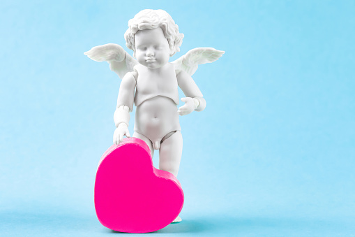 Retro cherub baby and pink heart on blue background. White cupid standing figure. Valentines day greeting card with cute small angel figurine