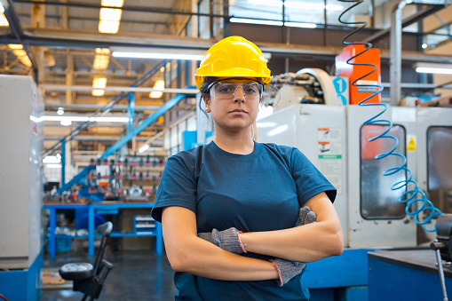 Portrait of confident female engineer wearing hardhat and protective eyewear standing with arms crossed in factory
