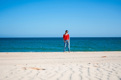 Young woman with red shirt and denim standing on beach sand with atlantic ocean in the back
