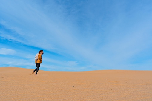 Asian woman walking up sandy sand dunes with blue cloudy sky above her.