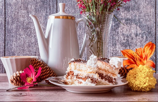 Positive summer still life with coffee pot, wildflowers in vase, slices of cake in plate and coffee mugs on dark wooden background.