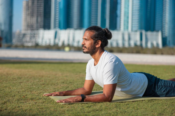 Man doing yoga on the lawn in public park in Abu Dhabi, lying on front and leaning on elbows in Cobra pose Bhujangasana, cityscape in the background, happiness and healthy lifestyle