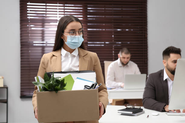 upset dismissed woman wearing protective mask carrying box with personal stuff in office - firing unemployment downsizing box imagens e fotografias de stock