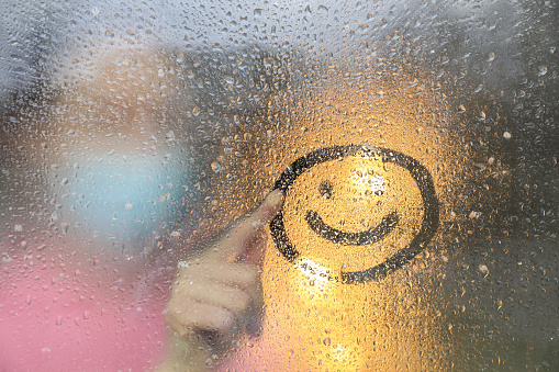 Woman drawing happy face on foggy window at rainy weather, closeup