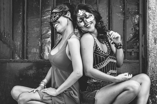 Black and white portrait of two beautiful and happy girls sitting against a green colored door. City girls far from the capital. They handle a carnival mask.