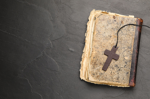 Wooden Christian cross and old Bible on black table, top view. Space for text