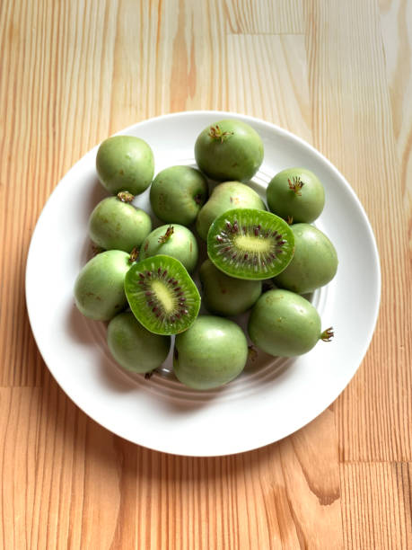 White plate with whole and halves of green kiwi or mini kiwi berries on a wooden background. Two halves of kiwi cut in half among whole berries. Vertical photo. View from above. White plate with whole and halves of green kiwi or mini kiwi berries on a wooden background. Two halves of kiwi cut in half among whole berries. Vertical photo. View from above. mini kiwi stock pictures, royalty-free photos & images