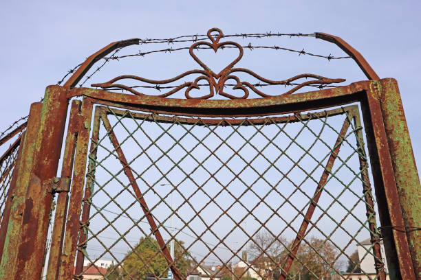 Ornamental silver-colored metal cross with curled scrollwork and bird spikes mounted on a gate, set against a curved ochre wall in direct sunlight.