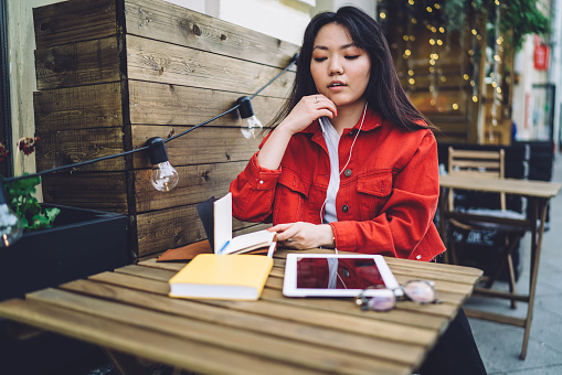 Focused Asian female freelancer in casual wear sitting at wooden table in outdoor cafe and listening to music in earphones while enjoying book
