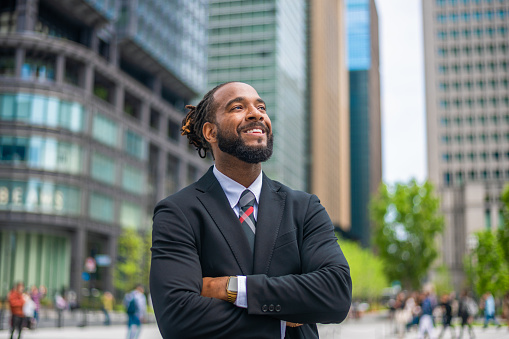 Waist up portrait of a successful mid adult black male entrepreneur. He is smiling and looking away with his arms crossed. He looks professional and is wearing formal businesswear. Defocused city buildings in the background.