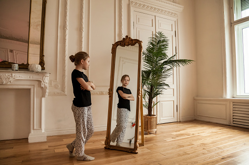 Alone cute teenager girl at mirror in living room at home, looking her. Sad lonely child stands and looks in his reflection in casual domestic clothes. Concept difficult  childhood. Copy text space