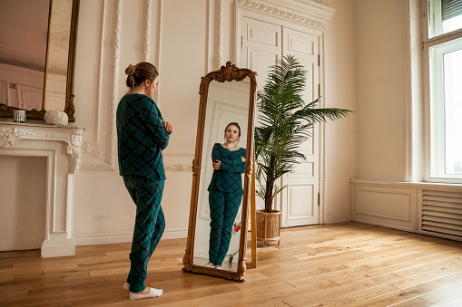 Lonely female looking at mirror in her reflection in living room at home. Alone middle aged adult woman standing and looks in mirror. Concept of adult domestic life and depression. Copy text space