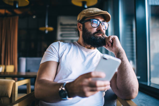 Pensive caucasian bearded male using mobile phone spending free time in coffee shop dreaming about plans and ideas, handsome mature man in eyewear pondering and resting with smartphone in cafeteria