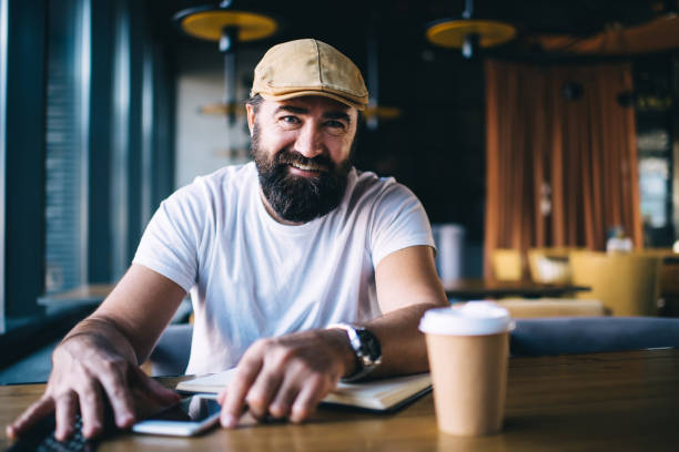 Cheerful hipster man smiling and looking at camera Bearded adult charismatic male sitting at table in cozy workspace with coffee and smartphone while smiling and looking at camera russian ethnicity stock pictures, royalty-free photos & images