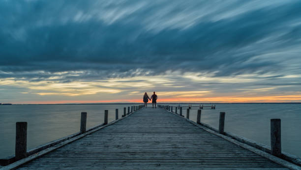 couple se tenant la main debout sur la jetée au crépuscule avec une rémanence au-dessus du lac - steinhuder meer photos et images de collection