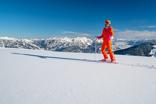 One female skier woman in colorful orange ski outfit with helmet standing alone in fresh snow high up in snow covered mountains on sunny skiing holiday vacation day