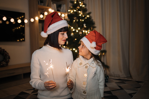 Smiling mom and little daughter in red santa hats, burning a sparkler celebrating the Christmas holiday in a cozy living room with a Christmas tree at indoor house.