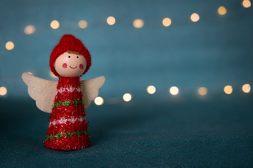 A cute red Christmas angel statuette with bokeh fairy lights on a blurred blue background. Christmas scene backdrop with decorative figurine and empty space for text