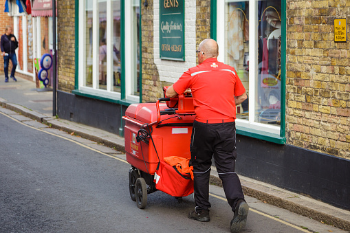 Sandwich, UK - 22 October, 2022: a senior male postal worker pushed his red Royal Mail trolley full of letters and packages on a street in the town of Sandwich in Kent, England.