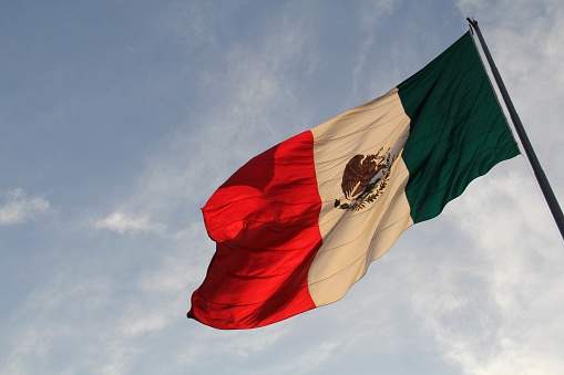 A large Mexican flag (Spanish: Bandera de México), lit with golden pre-sunset light, on a flag pole, with a blue sky, in the Zocalo, Mexico City