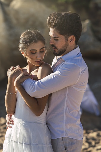A caucasian loving couple wearing white clothes and hugging in the beach during a wedding photoshoot