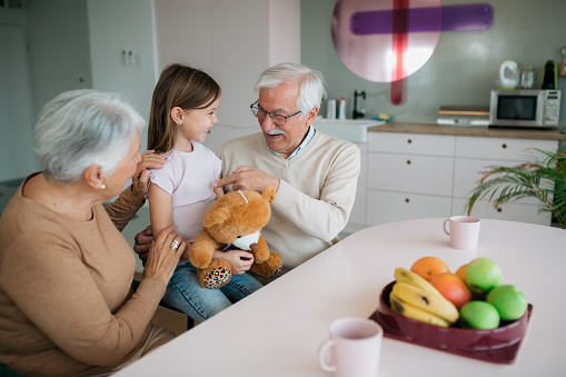Elderly grandparents having their granddaughter visiting them, surprising her by giving her a teddy bear plush toy as a present