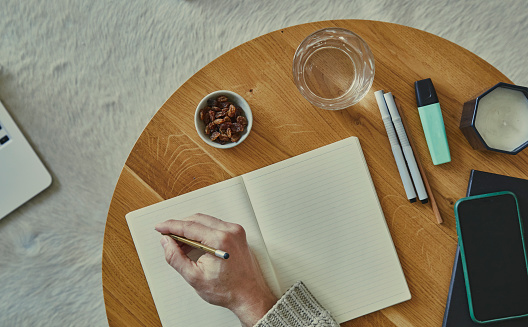 Mans hand holding a pen on a notebook, on a small home business desk, with a setting of pencils, mobile phone, marker, candle, a glass of water and a dry grapes in a small ceramic bowl
