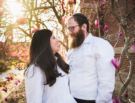 Outdoor portrait of mature Jewish couple smiling at each other; magnolia tree and sun flare in background