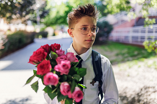 Teenage boy going to school on last day before vacations. Schoolboy is wearing white shirt with tie. The boy is holding a bouquet of flowers for the teacher.\nCanon R5