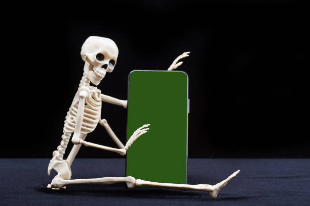 istock A seated skeleton is holding a smartphone with a green chromakey screen isolated on a black background 1437336115
