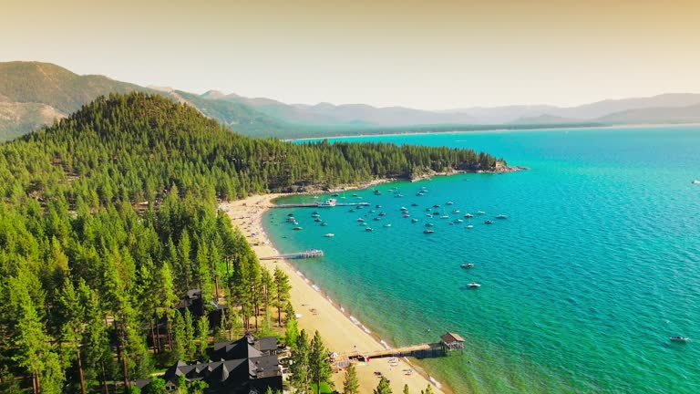 Lovely view of wonderful blue Lake Tahoe with boats at the shore. Sunny sandy beach and pine tree forest at waterfront. Hazy mountains at backdrop.