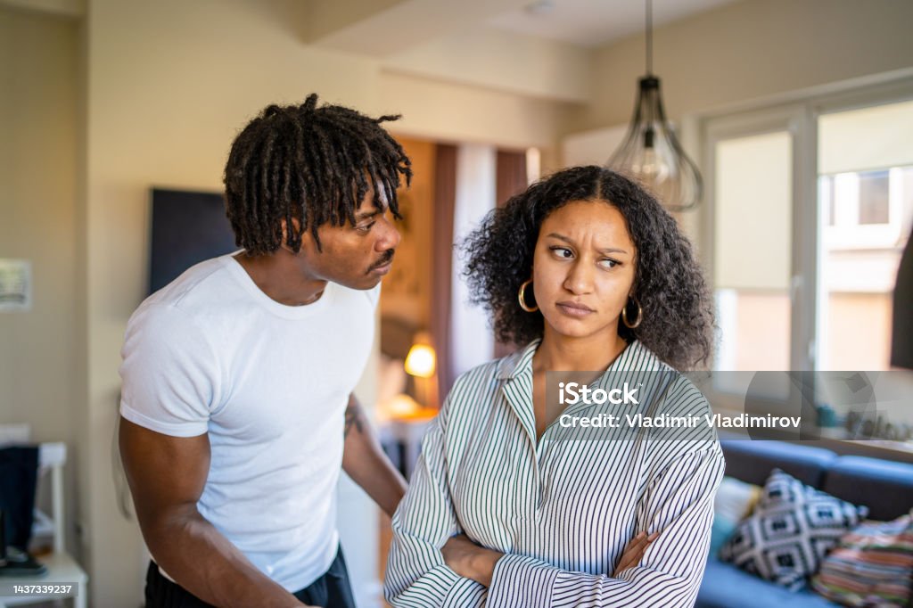 Frowning woman after argument with husband Woman is frowning and looking away from her boyfriend with her arms crossed, as he is looking worriedly into her face as they stand at home in their lounge Couple - Relationship Stock Photo