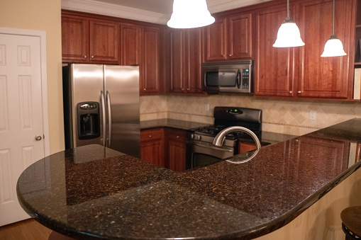A kitchen in dark wood finish with tile displays shining appliances in silver and black scheme. In focus in the foreground, a black granite countertop reflects the kitchen lights clearly