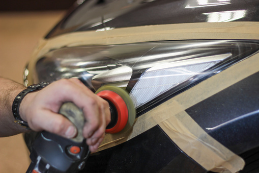 Restoration of the headlight glass using a grinding machine. A man polishes the optics of a car's headlights with a polishing machine at a service station. Hands working with a polishing machine.