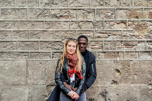 Portrait of smiling couple against stone building facade. Boyfriend embracing his girlfriend from behind.